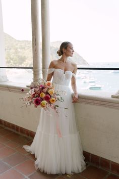 a woman in a white wedding dress standing on a balcony next to the ocean holding a bouquet of flowers