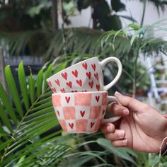 two coffee cups with hearts painted on them are held up in front of some plants