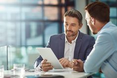 two men sitting at a table with a tablet in front of them and one is looking at the screen