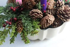 some pine cones are sitting in a white bowl with greenery and candles on it