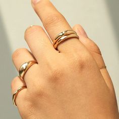 A hand with four weighty gold rings reaches towards a grey wall. The rings are all golden pieces that emulate dynamic motion. Modern Gold Stackable Dome Ring, Modern Rose Gold Dome Ring With Polished Finish, Modern Gold Rings For Anniversary, Modern Thick Band Midi Rings For Anniversary, Elegant Couple Rings With Thick Band, Elegant Rose Gold Dome Ring With Polished Finish, Modern Rose Gold Open Dome Ring, Shiny Finish Open Ring For Wedding, Classic Open Ring With Shiny Finish