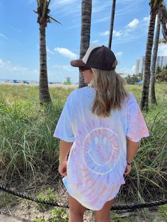 a woman standing in front of palm trees wearing a tie dye t - shirt and hat