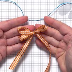 two hands holding an orange and white bow on top of a piece of paper with blue thread