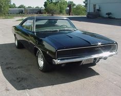 an old black muscle car parked in a parking lot next to a white building and trees