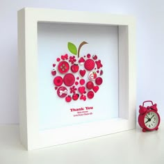 an apple made out of buttons in a white shadow box with a red alarm clock