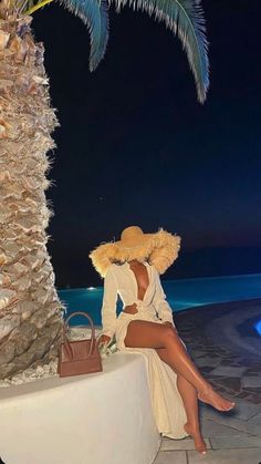 a woman sitting on a wall next to a palm tree at night wearing a straw hat