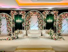 a wedding stage decorated with white furniture and floral arrangements on the wall, along with chandeliers