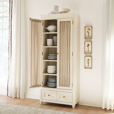 a white china cabinet with dishes on it in the corner of a room next to a window