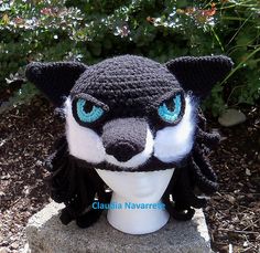 a crocheted black and white wolf head with blue eyes on a rock in front of some bushes