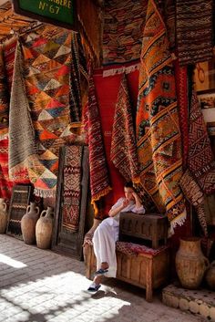 a person sitting on a bench in front of many rugs and vases hanging from the ceiling