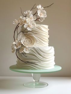 a three tiered cake with white frosting and flowers on the top is sitting on a glass plate