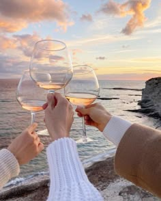 two people toasting wine glasses with the ocean and sky in the background at sunset