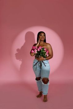 a woman in ripped jeans holding flowers and talking on a cell phone while standing against a pink background