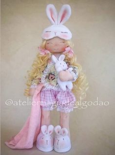 a doll with blonde hair holding a stuffed animal and wearing bunny ears, sitting on top of a pink blanket