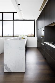 Elegant, sleek kitchen cabinetry in an inner-city penthouse kitchen. The dark wood tones with contemporary silver accents add a refined, dynamic edge to the space. The integrated double fridge is hidden behind this cabinetry. A double, stacked Miele oven sits on the wall and sleek, dark, hardwood floors complement the space. A gas cooktop is shown on the marble bench top and a long, marble island bench with a sink sits in front. Large, black-framed windows reveal the city views beyond. Dark Timber Floor Kitchen, Walnut Flooring Kitchen, Dark Floor Kitchen, Modern Kitchen Dark, Dark Hardwood Floors Kitchen, Black Floorboards, Suburban Family Home, Kitchen Marble Top, Dark Lower Cabinets