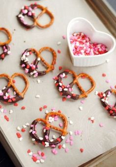 heart shaped pretzels and sprinkles are on a baking sheet next to a bowl of sprinkles