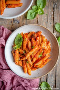 two bowls filled with pasta and basil on top of a wooden table next to a pink towel