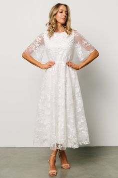 a woman wearing a white dress with sheer sleeves and flowers on the shoulders, standing in front of a wall