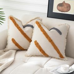 two decorative pillows on a white couch next to a coffee cup and framed pumpkins