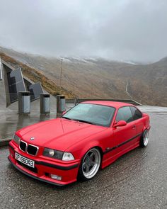 a red car parked on the side of a road near some hills and clouds in the background