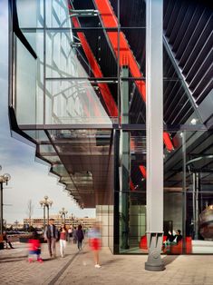 people are walking in front of a building with glass walls and red stairs on the side