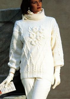 a woman in white sweater and pants holding a book while standing next to a stone wall