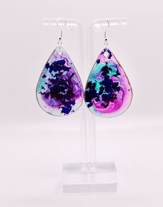 a pair of earrings with purple and blue paint on them, hanging from a clear acrylic stand