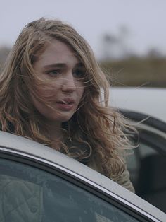 a woman with blonde hair standing in front of a car looking at the camera and making a face