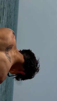 a man with tattoos on his chest standing next to the ocean and looking off into the distance