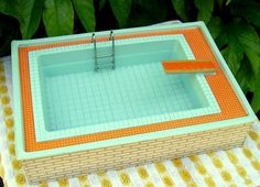 an orange and white table cloth with a miniature pool on it's edge next to a green bush