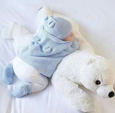 a white teddy bear laying next to a baby in a blue sweater and hat on a bed