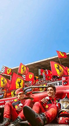 two men sitting on the ground in front of ferrari racing cars with flags behind them