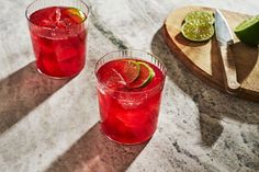 two glasses filled with red liquid sitting on top of a table next to sliced limes