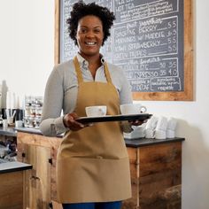 a woman in an apron holding a plate with a cup on it and smiling at the camera