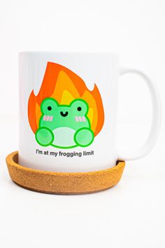 a white coffee mug with a green frog sitting on it's side in front of a fire