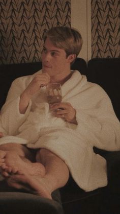 a man sitting on top of a black couch holding a drink in his hand and wearing a white bathrobe