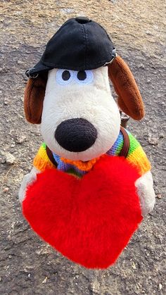 a stuffed dog wearing a hat and holding a red heart in its mouth on the ground