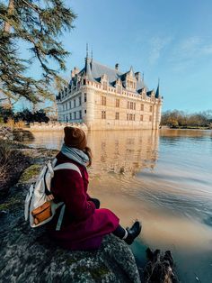 a woman sitting on top of a rock next to a body of water with a castle in the background