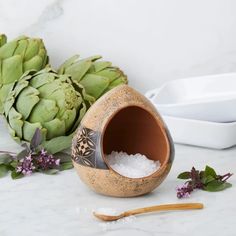 an artichoke and salt container sitting on a counter next to some flowers with spoons