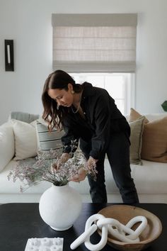 a woman arranging flowers in a white vase on a black table next to a couch