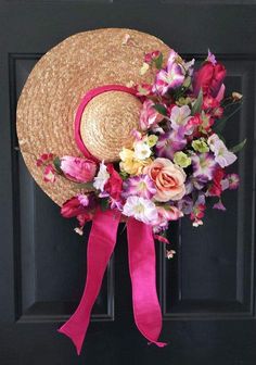 a straw hat with flowers and pink ribbon hanging on a black front door, next to a wreath