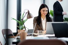 a woman sitting at a desk in front of a laptop computer with another person standing behind her