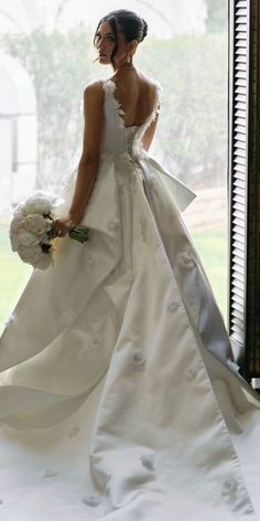 a woman in a white wedding dress holding a bouquet and looking out the window with her back to the camera