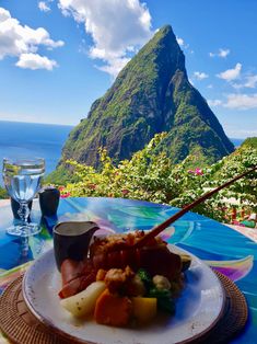 a plate with meat and vegetables on top of a table next to the ocean in front of a mountain