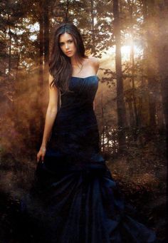 a woman in a black dress standing in the woods with her hand on her hip