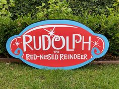 a red and blue sign that says rudolph the red - nosed reindeer in front of some bushes