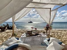 a table set up on the beach with white drapes over it and flowers in vases