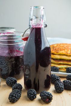 blackberry syrup in a glass bottle with blackberries around it on a wooden table next to pancakes