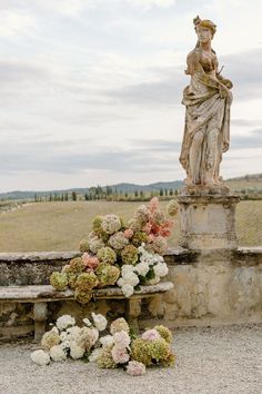 a statue sitting next to a bunch of flowers on top of a stone bench in front of a field