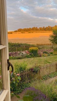 an open window looking out onto a garden and field in the distance with trees, bushes, and flowers